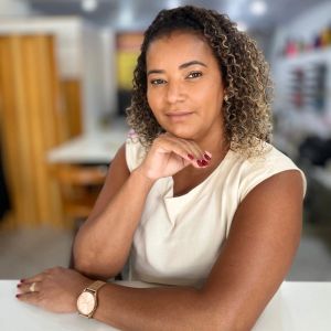 Mulher com expressão serena sentada em uma mesa de ateliê de costura, usando blusa clara e relógio dourado, com fundo desfocado de ambiente criativo e organizado